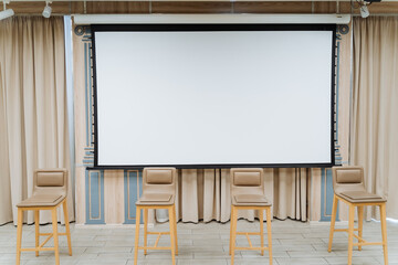 A white screen for showing the video hangs on the wall. Four chairs stand on the stage. Conference hall, business class for performances.