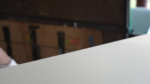 Selective focus of an Asian male factory worker's hand in a white fingerless working glove, picking up the cardboard on the pile put into a blurred  die cutting and creasing machine in a paper factory