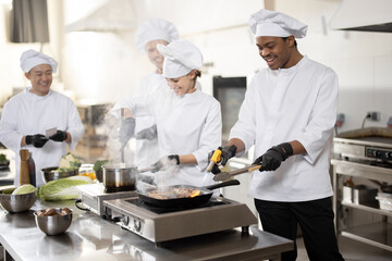 Fototapeta na wymiar Multiracial team of professional cooks in uniform preparing meals for a restaurant in the kitchen. Latin guy frying meat, european cooks making sauce and asian chef managing the process. Teamwork and