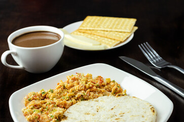 Colombian breakfast.Arepa (ground corn flour dough),egg with traditional sauce called hogado(stir-fried tomato and onion),with cup of chocolate,crackers and cheese,on background dark wood. Close up.