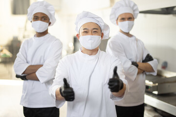 Portrait of three well-dressed chef cooks with different ethnicities standing together in restaurant kitchen. Asian chef showing OK sign, latin and european guys on background. Cooks wearing face