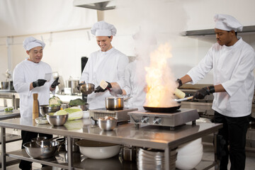 Multiracial team of professional cooks in uniform preparing meals for a restaurant in the kitchen. Latin guy burning pan, european cooks making sauce and asian chef managing the process. Teamwork and