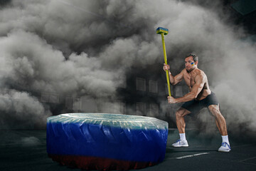 Muscular man with Ukrainian flag on cheeck hitting with hammer giant wheel with Russian flag on it...