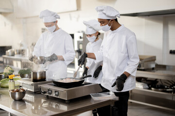 Multiracial team of cooks in uniform and face masks cooking meals for a restaurant in the kitchen. Concept of teamwork at restaurant during pandemic. Latin, Asian and European guys cooking together