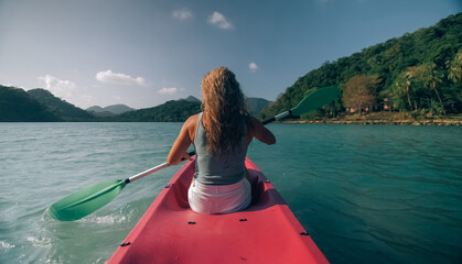 Long haired blonde woman with sunglasses rows bright pink canoe along sea bay water to beach with...