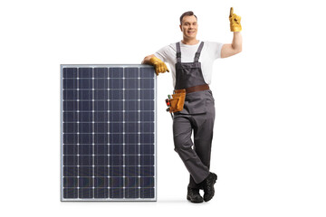 Full length portrait of a technician in a uniform leaning on a solar panel and pointing up