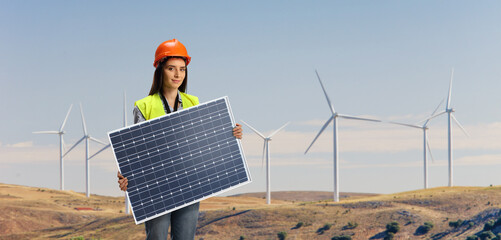 Female engineer holding a photovoltaic solar panel on a wind turbine field