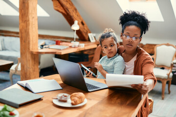 African American working mother using laptop and analyzing paperwork while daughter is sitting on her lap at home.