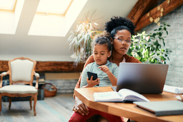 Small black girl uses cell phone while her mother is working on laptop at home.