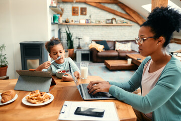 Fototapeta na wymiar Black little girl eats breakfast while her mother is working on laptop at dining table.
