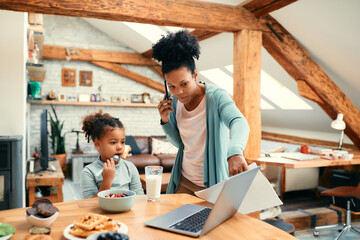 Black little girl eats while busy mother is giving her laptop to use during breakfast.