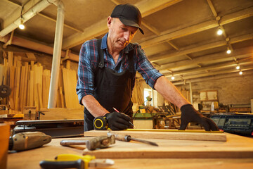 Portrait of a senior carpenter working with a wood, marking plank with a pencil and taking...