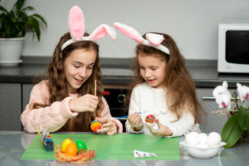 Obraz na płótnie Canvas Two girls in hare ears paint eggs. Easter. They are at home in the kitchen. Preparation for the holiday.