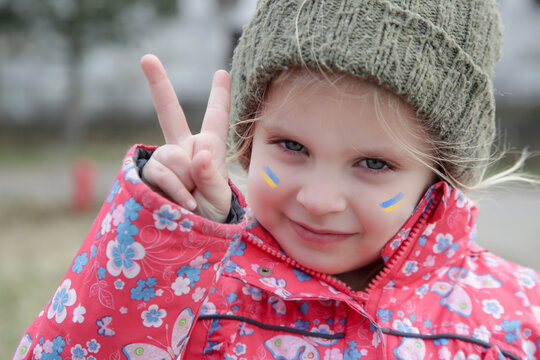 Portrait of little girl with Ukraine flag painted on her face showing Victory sign. Refugees, war crisis, humanitarian disaster concept.