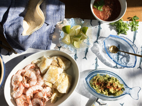 summer Seafood supper table spread to make fish tacos, prawns, guacamole, lime, tortilla, salsa on blue and white fish tablecloth in the sun
