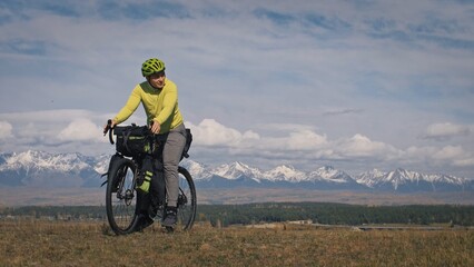 Naklejka na ściany i meble The man travel on mixed terrain cycle touring with bikepacking. The traveler journey with bicycle bags. Sport tourism bikepacking, bike, sportswear in green black colors. Mountain snow capped.