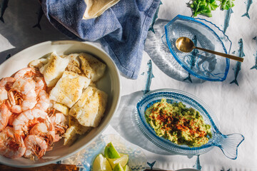 summer Seafood supper table spread to make fish tacos, prawns, guacamole, lime, tortilla, salsa on blue and white fish tablecloth in the sun