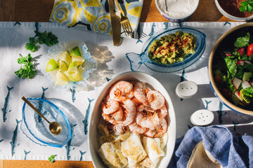 summer Seafood supper table spread to make fish tacos, prawns, guacamole, lime, tortilla, salsa on blue and white fish tablecloth in the sun