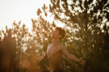 Young man doing yoga at the sunrise on a top of a mountain next the trees over the rocks with arm wide open