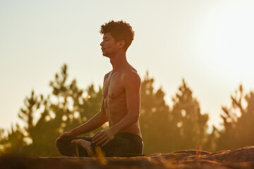 Young man doing yoga at the sunrise on a top of a mountain next the trees over the rocks