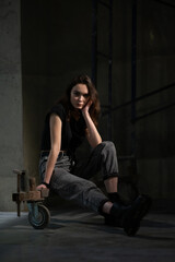 Fototapeta na wymiar A beautiful young girl sits on scaffolding in a beam of light. Dressed in jeans and black boots
