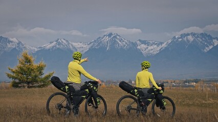 The man and woman travel on mixed terrain cycle touring with bikepacking. The two people journey with bicycle bags. Sport bikepacking, bike, sportswear in green black colors. Mountain snow capped.