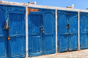 Warehouses closed with blue wooden gates in the old fishing harbor of the Essaouira on a sunny summer day. Morocco.