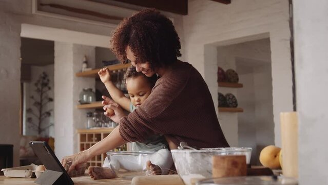 Biracial mother baking with her young daughter in a modern-styled kitchen, cracking eggs in bowl to whisk.