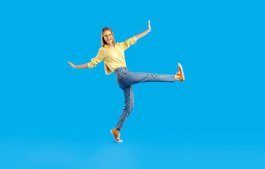 Fototapeta na wymiar Funny cheerful pretty girl in comfortable casual clothes having fun in studio. Happy slim young woman in shirt, jeans, eyeglasses and canvas sneakers walking isolated on solid blue background