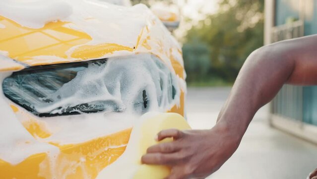 Closeup cropped image of hand of young African man with yellow sponge washing headlight of his car at a self-serve car wash outdoors. Luxury car covered by foam. Foaming after rubbing the car