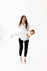 Happy harmonious family indoors in studio. Attractive young mother wearing high heels and white blouse throws the baby girl up, laughs and plays together on the white background