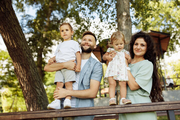 Lovely parents with little kids at the park, caring father hold in arms joyful son, cute daughter sitting in mothers hands, enjoy happy family moments, weekend outdoors concept