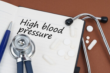 On a brown surface lie pills, a pen, a stethoscope and a notebook with the inscription - High blood...