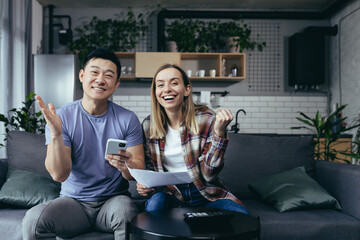Obraz premium homework paper, multiracial couple, man and woman, pay bills by phone and app, sit at home on the couch, happy and smiling