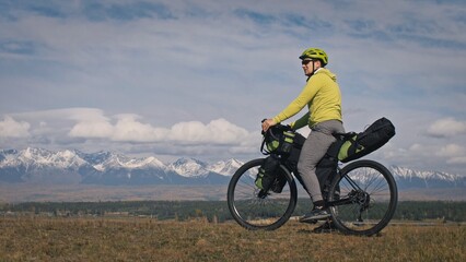 Fototapeta premium The man travel on mixed terrain cycle touring with bikepacking. The traveler journey with bicycle bags. Sport tourism bikepacking, bike, sportswear in green black colors. Mountain snow capped.