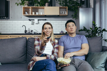 Young couple in love man and woman watching TV together at home, rejoicing and eating popcorn, happy multiracial family in a new apartment
