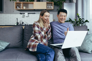 Happy young asian couple bonding laughing using laptop sit at table home