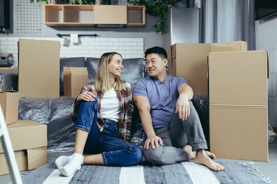 Young multiracial couple Asian man and woman, in a newly rented apartment, hugging sitting on the floor of the house, cardboard boxes unpacked - Powered by Adobe