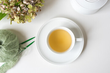 High angle closeup of herbal tea in white cup and saucer on table with knitting and dried hydrangeas (selective focus)