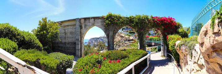 a panoramic of The Dana Point Arches with lush green plants and red flowers down a staircase with a white hand rail with gray homes along the mountain and blue sky at Dana Point Bluff Top Trail - obrazy, fototapety, plakaty