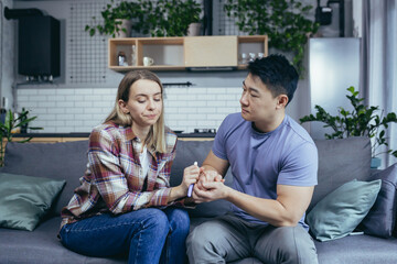 Man Asian supports wife, disappointing pregnancy test result, young multiracial couple sitting on sofa, frustrated