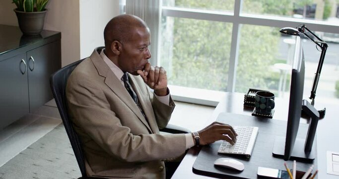 Side profile view of a happy corporate black business man typing on a desktop computer while talking on a headset near a large office window. 35mm Medium Closeup Tripod Shot 4K.