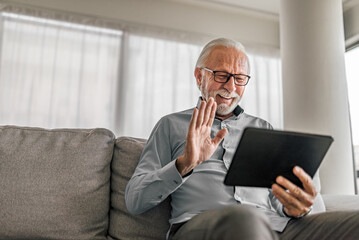 Cheerful senior man having video call on tablet sitting on the couch at home elderly man wearing eyeglasses staying in touch with friends and family using online video call connecting with people