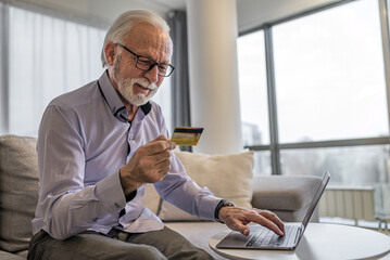 Senior good-looking handsome man sitting at his home or office using laptop computer and credit card for online shopping web paying, buying presents for friends and family