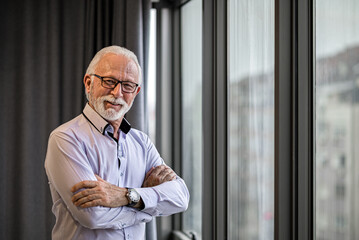 Portrait of smiling cheerful satisfied mature white bearded man with eyeglasses standing next to large window in his office arms crossed looking at camera.