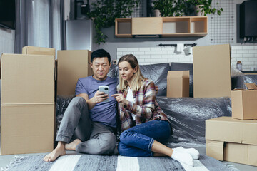 A young family in a new apartment among the cardboard boxes sitting on the floor and happily choosing new furniture online store, use the phone app for shopping