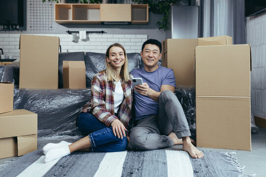 A young family in a new apartment among the cardboard boxes sitting on the floor and happily choosing new furniture online store, use the phone app for shopping - Powered by Adobe