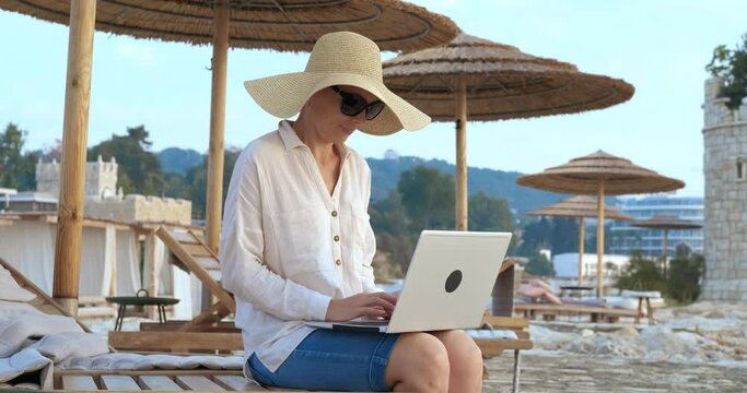 Working freelancer on traveling. A woman in straw hat relax on the bech and work on remote online on her notebook.