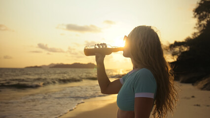 Sexy hot silhouette woman drinking whiskey with orange juice on beach. Rays gold sunset flare through glass. Woman in blue swimsuit, sunglasses. Concept happy summer holiday, alcohol drink, beverages