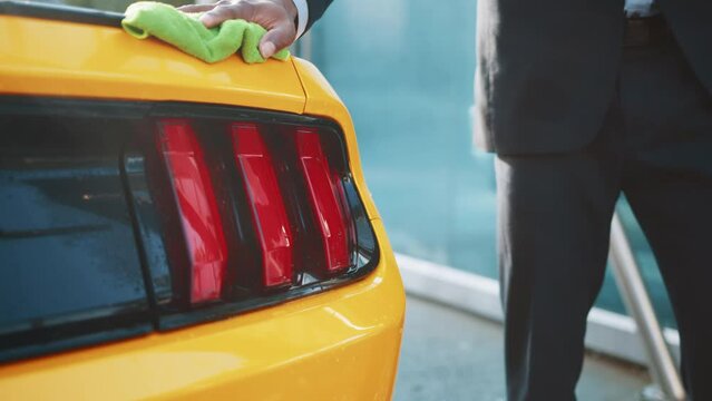 Closeup cropped image of hand of young African businessman, washing and cleaning yellow sport car with microfiber cloth, wiping hood and headlights. Car wash outdoors concept.